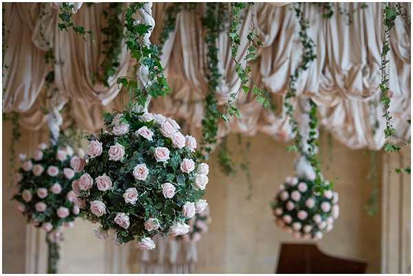 A close-up detail shot of suspended floral orbs made from blush pink roses and dense green foliage, hanging from the ceiling of what appears to be an indoor ballroom or reception hall. Trailing ivy or similar vines drape alongside the spheres, and the background features gathered champagne or ivory fabric swags decorating the walls. The decor palette combines blush pink, green, and warm ivory tones in a classic, romantic styling. Two full floral balls are visible in the foreground with a third partially visible to the right.