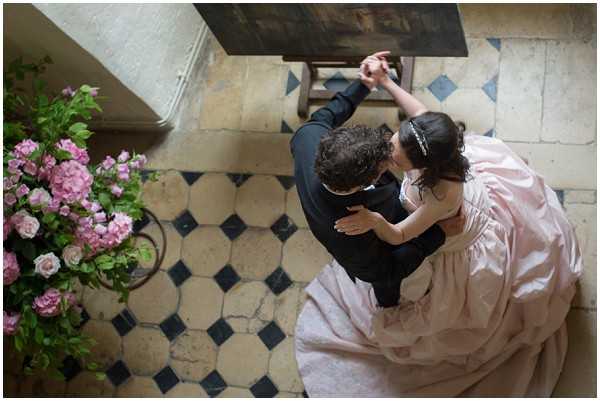 An aerial overhead shot of a couple sharing a first dance on a classic black-and-cream geometric tiled floor, likely inside a French chateau. The groom is dressed in a dark suit and holds the bride's hand aloft while she wraps her other arm around him. The bride wears a full-skirted blush pink ballgown with a low back, and her dark hair is accessorized with a crystal or pearl headband. To the left, a large floral arrangement in a basket features pink hydrangeas and pale pink spray roses. The composition captures the couple from directly above, emphasizing the sweeping skirt of the gown and the ornate tile floor beneath them.