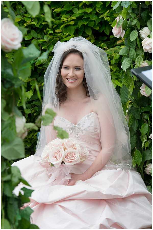 A bridal portrait taken outdoors, with the bride seated against a lush green climbing vine wall dotted with blush and soft pink roses. The bride wears a blush pink strapless ballgown with a crystal and beaded sweetheart bodice, paired with a two-tier elbow-length veil. She holds a compact round bouquet composed entirely of soft blush pink roses. The composition is a close-up portrait framing the bride from approximately the waist up, with climbing roses visible in the foreground and background adding depth to the shot.