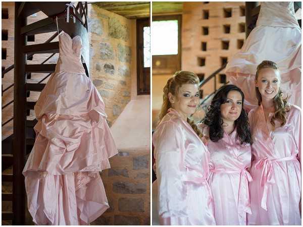 A split image showing two getting-ready scenes inside a rustic stone building, likely a chateau or barn with exposed stone walls and a dark wooden spiral staircase. The left panel is a detail shot of a blush pink strapless ball gown with a ruched and layered skirt, hanging from the spiral staircase railing. The right panel is a portrait of three women — likely the bride and two bridesmaids — posing together, all wearing matching light pink satin robes with tie waists; their hair and makeup are already done, suggesting they are mid-preparation. The overall palette centers on blush and light pink, and the setting has a classic rustic character.