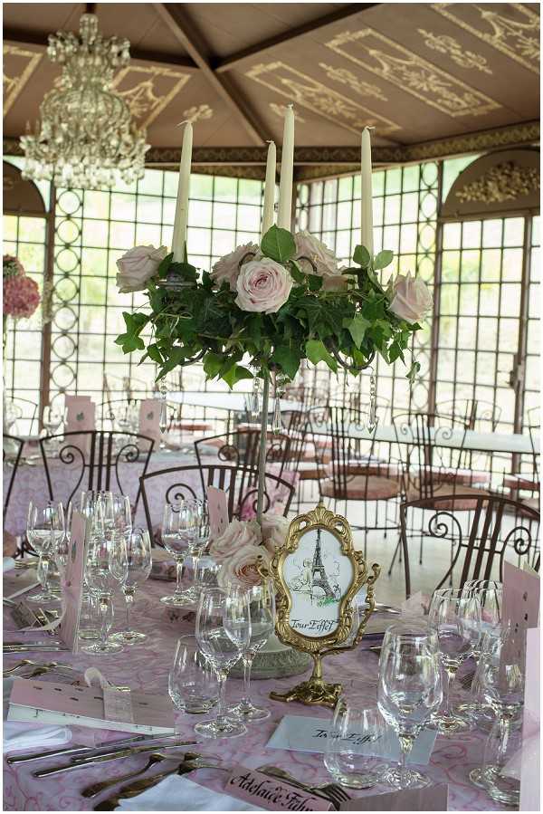 A close-up detail shot of a wedding reception table setting inside a glass-paneled pavilion or orangery with an ornate painted ceiling and a crystal chandelier overhead. The round table is dressed in a blush pink damask-patterned tablecloth and set with multiple crystal wine glasses, gold cutlery, and white napkins. The tall centerpiece features a candelabra topped with blush pink roses and trailing green ivy, holding five white taper candles. A small gold ornate frame displaying an Eiffel Tower illustration serves as a Paris-themed table name card reading 'Tour Eiffel,' establishing a French capital theme for the wedding. Additional round tables with wrought iron chairs are visible in the background, along with what appears to be a pink floral arrangement. The overall decor palette combines blush pink, gold, and green in a classic French-inspired style.