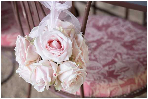 Close-up detail shot of a round pomander ball of blush pink roses tied with a sheer pale pink ribbon, hanging from the back of a dark wood chair with a red and white toile de Jouy cushion seat. The floral arrangement is tightly packed with fully open roses in a uniform soft blush tone. The romantic, classic French-inspired styling is reflected in the toile fabric pattern visible in the soft-focus background.