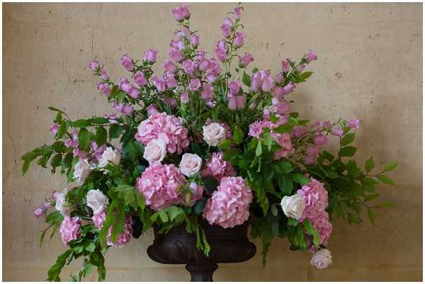 A close-up detail shot of a large floral arrangement displayed in a dark bronze/iron urn pedestal vase. The arrangement features pink hydrangeas, blush pink roses, and tall mauve-purple delphinium spires, with abundant green fern fronds and foliage trailing outward on both sides. The overall color palette is soft pink and blush with lush greenery, styled in a full, asymmetric garden-style arrangement. The background is a pale, textured plaster wall.