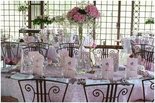 A reception tablescape detail shot taken indoors in what appears to be a glass-walled or windowed venue with decorative iron lattice panels. The round tables are dressed in pink patterned tablecloths and set with crystal glassware, silverware, and pink folded menu cards decorated with a small blue butterfly motif. Place cards are visible along the table edge. The centerpiece is a tall clear vase filled with pink hydrangeas and baby's breath. The chairs are dark wrought-iron with scrolled backs. The overall decor palette is soft pink and white with romantic, classic styling throughout the room.