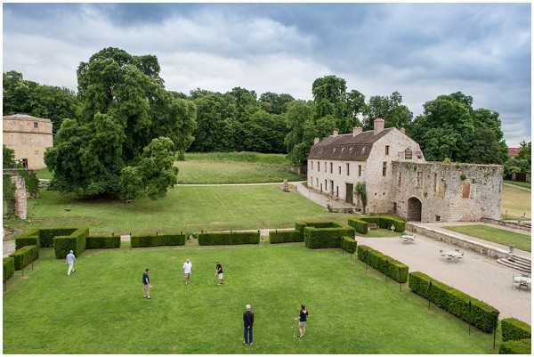 An elevated wide shot looking down over the manicured grounds of a French stone chateau or manor property, showing a large formal lawn bordered by neatly trimmed box hedges arranged in a geometric pattern. Six people in casual attire are scattered across the lawn, appearing to play croquet or a similar lawn game. The property features a substantial medieval-style stone building with a ruined archway section to the right, and a gravel terrace with white garden furniture visible in the background. The setting suggests a pre-wedding or cocktail hour activity during a cloudy day. Potential venue feature image.