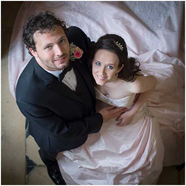 A couple portrait shot from directly above, with both the bride and groom looking up at the camera. The groom wears a dark charcoal suit with a white shirt and a boutonniere featuring a pink rose with baby's breath. The bride wears a strapless blush pink ballgown with a beaded silver bodice, and her dark hair is accessorized with small white flowers. The voluminous skirt of the dress fans out around the couple, filling much of the frame. The groom has his arms around the bride from behind as they sit together on what appears to be a stone floor. The composition is an overhead portrait shot that uses the spread of the dress as a natural framing device.