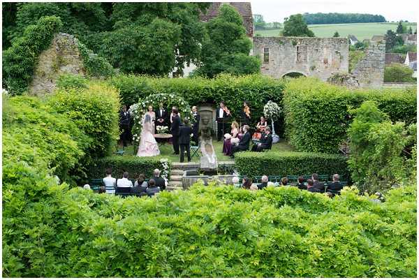 An outdoor wedding ceremony photographed from an elevated wide-angle perspective, showing a formal garden setting with neatly trimmed tall hedgerows and a stone statue or fountain as a focal point at the altar area. The couple stands at the front alongside what appears to be an officiant and a small string quartet or chamber ensemble seated to one side, with two white floral arrangements flanking the ceremony space. Approximately 30-40 guests are seated in rows on garden chairs facing the altar. The bride appears to be wearing a pale pink gown. Historic stone ruins are visible in the background, suggesting a French chateau or abbey grounds. The decor is classic and understated, with white florals against the green hedging. Potential venue feature image.