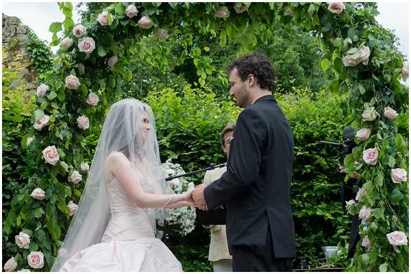 An outdoor wedding ceremony showing a bride and groom exchanging vows while holding hands, with an officiant visible in the background. The ceremony takes place beneath a large circular floral arch decorated with lush green foliage and blush pink roses. The bride wears a strapless white ballgown with a long veil and holds a small white and blush bouquet, while the groom is dressed in a black suit. The setting appears to be a garden with dense greenery. The shot is a medium portrait-style image taken from a slight side angle, capturing both the couple and the decorative arch.
