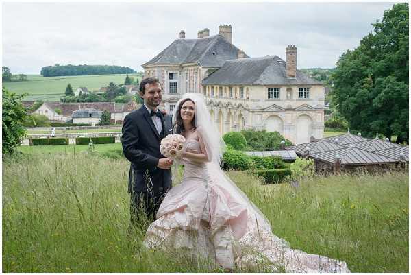 A couple poses together in a wide outdoor portrait shot on the grounds of a French chateau, with the historic stone manor house clearly visible in the background. The bride wears a voluminous blush pink ballgown with a cathedral-length train spread across the tall grass, paired with a white veil and a rounded bouquet of blush and cream flowers. The groom is dressed in a dark navy tuxedo with a black bow tie. The setting is a sprawling lawn with overgrown grass in the foreground and the château's classical French architecture — including ornate stonework and tall chimneys — prominently featured behind them. Potential venue feature image.