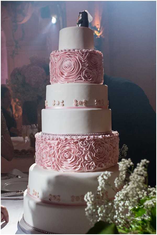 Close-up detail shot of a five-tier wedding cake displayed at an indoor reception. The cake alternates between smooth white fondant tiers and dusty pink rosette-textured tiers, with pearl bead detailing along the borders of each layer. The top is finished with a figurine cake topper depicting a bride and groom. White gypsophila (baby's breath) blooms are visible in the foreground, and cake serving utensils can be seen on a plate nearby. The background shows warm amber uplighting in what appears to be a reception venue, with a guest partially visible. The overall decor palette is white and dusty pink with a classic, formal style.