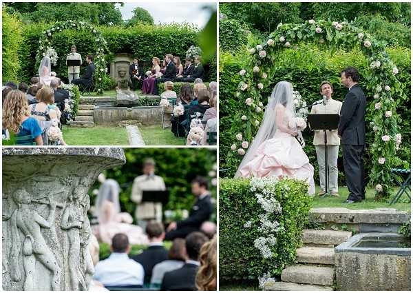 A three-image collage of an outdoor garden wedding ceremony. The bride wears a full-skirted blush pink ball gown with a long cathedral veil, and stands with the groom in a dark suit beneath a large floral arch decorated with white and blush roses and lush green foliage. An officiant reads from a lectern beside them. The ceremony takes place on stone steps surrounded by dense hedging and mature garden planting, with a decorative stone fountain or garden sculpture visible in the foreground of one image. Approximately 30-40 guests are seated in rows of chairs facing the altar. The bottom-left image is a close-up detail shot of the carved stone garden sculpture with the ceremony blurred softly in the background. The top-left image provides a wide shot of the full ceremony setup showing the floral arch, bridal party in dark attire with at least one bridesmaid in a dark plum dress, and seated guests. The styling is classic and formal with a soft romantic color palette of blush and white florals.