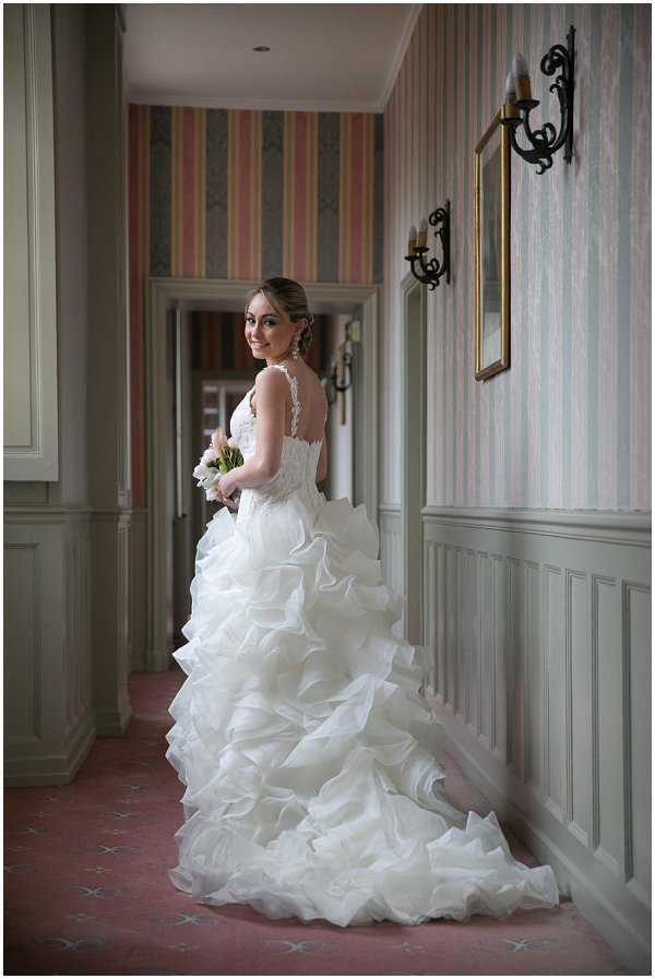 A bridal portrait taken indoors in a corridor or hallway of what appears to be a classic French chateau or manor house. The bride stands three-quarter turned toward the camera, looking back over her shoulder with a smile, wearing a white ball gown with a lace illusion back bodice and a voluminous tiered ruffle skirt with a long train. She holds a small bouquet of pale pink and white flowers, likely tulips or roses. The hallway features grey-painted wood panelling on the lower walls, striped wallpaper in muted pink and grey tones on the upper walls, a gold-framed mirror, and a wrought-iron candle wall sconce. The floor is covered in pink carpeting with a subtle geometric motif. The composition is a full-length portrait shot with shallow depth of field, with a second doorway visible in the background.
