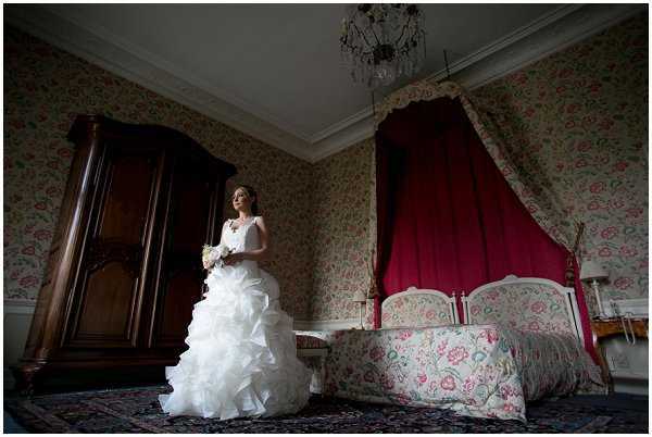 A bridal portrait taken indoors in what appears to be a period-style chateau bedroom. The bride stands alone near a large dark wooden armoire, wearing a white strapless ball gown with a heavily ruffled and layered skirt, holding a small white bouquet. The room features floral-patterned wallpaper in pink and green tones, a four-poster bed with deep crimson curtains and a matching floral bedspread, an ornate crystal chandelier overhead, and a patterned area rug on the floor. The image is shot as a wide portrait with dramatic natural light coming from the left side, leaving parts of the room in shadow and creating strong contrast between the bright white dress and the darker surroundings.