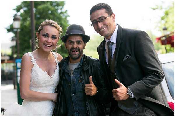 A candid portrait of three people posing together outdoors, likely during a wedding day. The bride wears a white lace V-neck dress with an updo hairstyle, standing to the left, while the groom on the right is dressed in a charcoal grey suit with a light grey tie and pocket square. Between them stands a guest or entertainer wearing a grey fedora hat, denim jacket, and leather jacket, giving two thumbs up with a wide smile. The setting appears to be a street or outdoor area with trees visible in the background, and the shot is a close-up portrait-style composition.