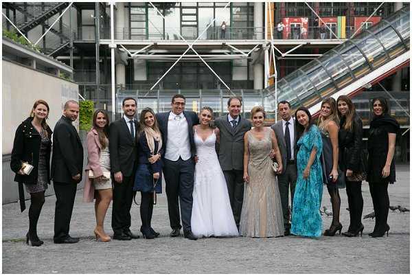 A wedding group portrait taken outdoors in front of the Centre Pompidou in Paris, with the building's distinctive exposed structural framework, escalator tubes, and industrial facade clearly visible in the background. The group of approximately 13 people is posed on the plaza, centered around the bride in a white strapless lace ball gown and the groom in a dark suit with a white vest and bow tie. Guests are dressed in formal attire including a teal/turquoise floor-length gown, a gold-toned midi dress, a pink blazer with a short skirt, and a navy dress, with most other guests in black. Wide group shot taken at ground level in overcast daylight.