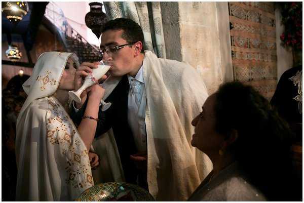 A couple shares a ceremonial drink from a decorative cup during what appears to be a Jewish wedding ceremony, likely the kiddush or the sharing of wine under the chuppah. The bride wears a white hooded cape or veil with gold embroidered detailing, while the groom wears a dark suit with a white tallit (prayer shawl) draped over his shoulders and glasses. A female guest or officiant is visible in the foreground. The setting is indoors with ornate brick or carved stone walls, hanging lanterns with intricate metalwork, and warm ambient lighting, suggesting a historic or traditionally decorated venue. The shot is a close-up portrait capturing an intimate ceremonial moment.