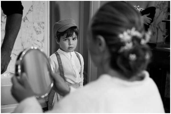 A black-and-white getting-ready scene featuring two young children, likely a page boy and a flower girl. The boy, positioned in the center of the frame, wears a flat cap, white shirt, and suspenders, and looks directly toward the camera with a serious expression. In the foreground, the flower girl is seen from behind, her hair styled in an updo decorated with small white floral pins, and she holds a round hand mirror. The setting appears to be an indoor preparation room with a marble surface visible in the background. The image is a candid medium shot with soft contrast, capturing an unguarded moment between the two children during wedding preparations.
