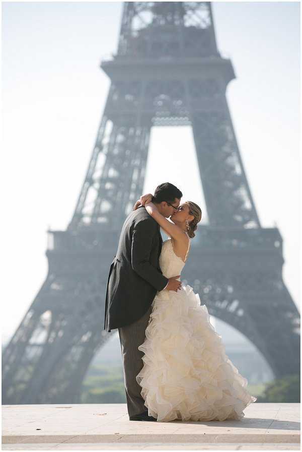 A couple portrait shot outdoors at the Trocadéro esplanade in Paris, with the Eiffel Tower deliberately blurred in the background through shallow depth of field. The bride wears a strapless ivory ballgown with a heavily ruffled and layered skirt, and the groom wears a dark charcoal grey suit with pinstripe trousers. The two are kissing, with the bride's arm around the groom's neck, in a dip-style pose. The composition is a medium full-length portrait framing both figures centrally against the iconic landmark backdrop, shot in natural daytime light with a slightly hazy atmosphere.