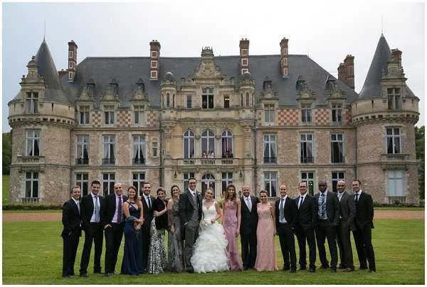 A wide-shot group portrait of approximately 20 people posed on the lawn in front of a large French Renaissance-style chateau with stone facades, ornate dormer windows, checkerboard brick detailing, and corner turrets with slate roofs. The bride stands near the center wearing a voluminous white ruffled ballgown and holding a white bouquet, flanked by the groom in a grey suit. The group includes men in dark suits and ties and women in a variety of formal dresses including a dusky pink floor-length gown, a navy blue gown, and a floral-print dress. The overall styling is classic and formal. Potential venue feature image.