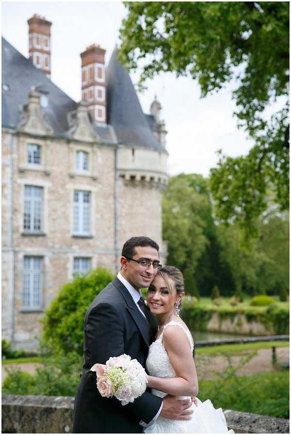 A couple portrait taken outdoors on the grounds of a French chateau, with the stone and brick Renaissance-style castle visible and slightly out of focus in the background. The groom, wearing a dark navy suit with a light blue dress shirt, stands behind the bride with his arms around her waist. The bride wears a sleeveless lace-bodice white gown and holds a bouquet of blush pink roses and white blooms with small filler flowers. The composition is a medium portrait shot with the couple centered in the foreground and the chateau's distinctive conical turret and red brick chimneys framing the background.