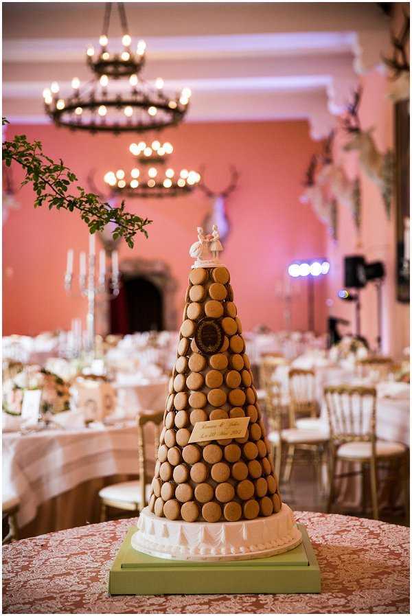 Close-up detail shot of a traditional French macaron tower (pièce montée) centerpiece at a wedding reception, displayed on a sage green base with a white decorative cake board and topped with a classic bride-and-groom ceramic figurine topper. The tower features rows of uniform tan/caramel-colored macarons with a personalized gold plaque bearing the couple's names and wedding date. The tower is placed on a table with a dusty rose damask-patterned linen. In the background, the reception room features salmon-pink walls, mounted stag antler decor, a wrought-iron chandelier with warm bulb lighting, tall candelabras with white tapers, and round guest tables set with gold Chiavari chairs and neutral linens, indicating a classic French château interior with a warm, candlelit reception atmosphere.