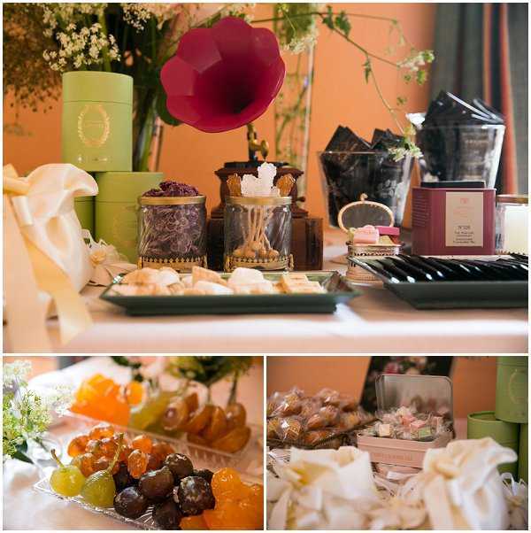 Detail shots of a wedding sweet table or gift/favor display, shown in a three-image collage. The top image shows a styled table against a peach-toned wall featuring green cylindrical gift boxes with gold lettering, glass jars with gold-trimmed lids containing sugar crystals and dried botanicals, a bold crimson flower-shaped decorative object, dark gift bags, a burgundy boxed item, a small ornate mirror, and a tray of macarons and petit fours. The bottom-left image shows a platter of candied and dried fruits including glazed cherries, orange slices, and chocolate-coated pieces in amber, orange, and dark brown tones. The bottom-right image shows cellophane-wrapped individual pastries or cannoli arranged in a tin, alongside pink and green favor boxes and ivory ribbon. The overall styling palette combines deep burgundy, sage green, and gold accents, suggesting a classic French confectionery or tea-inspired favor table. Close-up and detail composition throughout.