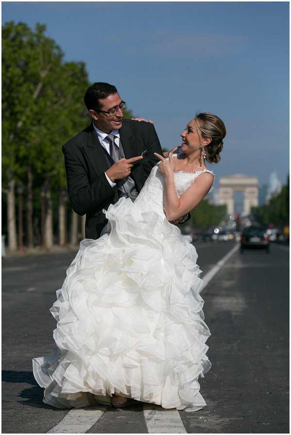 A couple poses playfully on the Avenue des Champs-Élysées in Paris, with the Arc de Triomphe clearly visible in the background. The bride wears a white ballgown with a heavily layered, ruffled organza skirt and a lace bodice with cap sleeves, her hair styled in an updo with drop earrings; the groom wears a dark charcoal suit with a grey tie and glasses. Both are laughing and pointing at each other in a candid, lighthearted moment mid-street, with passing cars visible in the background. The shot is a full-length portrait taken at street level with a shallow depth of field that keeps the couple in sharp focus against the blurred Parisian boulevard.