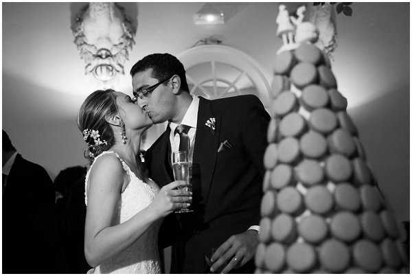 Black-and-white portrait of the bride and groom sharing a kiss during the reception, likely at the wedding cake cutting moment. The bride wears a lace-detailed dress with floral hair accessories and holds a champagne flute, while the groom wears a dark suit with a boutonniere and glasses. In the foreground, a pièce montée (traditional French macaron tower) is partially visible, and ornate plasterwork decor is visible on the wall behind the couple. The image has high contrast with bright uplighting illuminating the couple against a darker background.