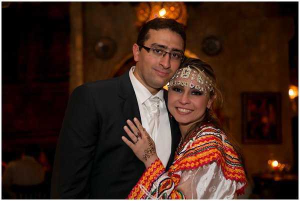 A couple poses together in a close portrait shot during what appears to be a wedding reception with North African or Middle Eastern cultural styling. The groom wears a dark suit with a white dress shirt and white tie, while the bride is dressed in a traditional embroidered garment featuring bold red, orange, green, and white geometric patterns. She wears an ornate silver and white beaded headpiece, dramatic eye makeup, and has henna designs visible on her hand. The indoor venue features warm amber lighting from decorative Moorish-style pendant lanterns and ornamental wall details visible in the background, creating a richly styled atmosphere. Candles and additional warm light sources are faintly visible in the far background.
