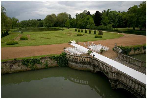 An aerial wide shot of an outdoor wedding ceremony setup at a French château estate, viewed from an elevated vantage point. A white aisle runner extends across a stone balustrade bridge over a moat or ornamental canal, leading to rows of white chairs arranged in a ceremony configuration on a gravel terrace. The formal French garden beyond features manicured lawns, clipped topiary cones, and geometric hedging stretching into the distance. One person is visible on the bridge, appearing to make final preparations. Potential venue feature image.