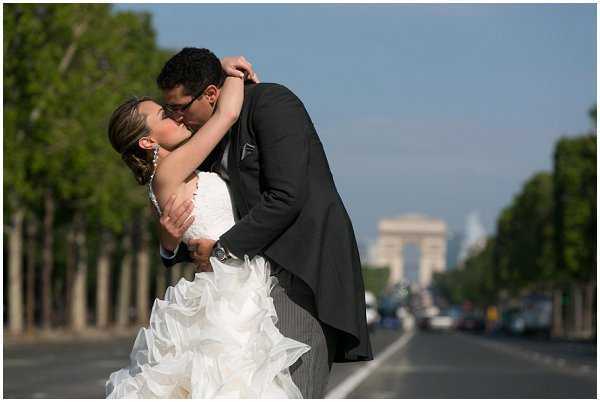 A couple shares a dip kiss during a wedding portrait session on the Champs-Élysées in Paris, with the Arc de Triomphe visible in the background. The bride wears a white strapless ball gown with a heavily ruffled, layered skirt and statement earrings, while the groom is dressed in a dark charcoal suit with pinstripe trousers. The shot is taken from street level with the boulevard stretching into the distance, creating a strong sense of depth with light traffic visible. The composition is a mid-range portrait with a shallow depth of field that keeps the couple sharp against the softly blurred Parisian backdrop.