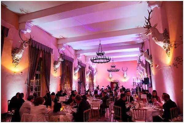 A wedding reception dinner is underway inside a grand ballroom or hunting-lodge-style salon, with approximately 60–80 guests seated at round tables covered in white linens. The room is decorated with multiple white-painted stag head mounts mounted on the walls, wrought-iron chandelier with candle-style lights hanging from the ceiling, and tall candelabras on the tables. The venue is lit with warm amber uplighting on the side walls and vivid pink-purple wash lighting on the back wall, creating a dramatic, moody atmosphere. The decor style blends classic French chateau hunting-room architecture with theatrical colored lighting. The shot is a wide-angle view from the back of the room looking toward the far wall. Potential venue feature image.