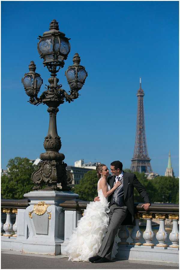 A couple poses for wedding portraits on the Pont Alexandre III bridge in Paris, with the Eiffel Tower visible in the background. The bride wears a strapless white ballgown with a heavily ruffled and layered skirt, while the groom is dressed in a charcoal grey suit with a tie; the two are seated and standing against the ornate white and gold balustrade of the bridge. A large decorative cast-iron candelabra lamppost with blue glass globes dominates the left foreground, and the bridge's signature gold-accented shell motifs are visible on the stone railing. The shot is a medium-wide portrait taken in natural daylight, with classic Parisian architecture framing the couple.