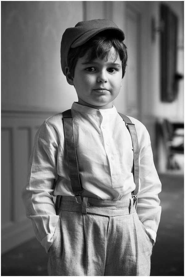 Black-and-white close-up portrait of a young boy dressed as a page boy or ring bearer, standing indoors with his hands in his pockets and looking directly at the camera. He wears a linen collarless shirt, matching high-waisted linen trousers, leather suspenders, and a flat cap, styled in a classic vintage-inspired look. The background is softly blurred, showing panelled walls and what appears to be an interior doorway, with good contrast between the light tones of his outfit and the mid-grey background. The composition is a tight mid-length portrait shot with shallow depth of field.