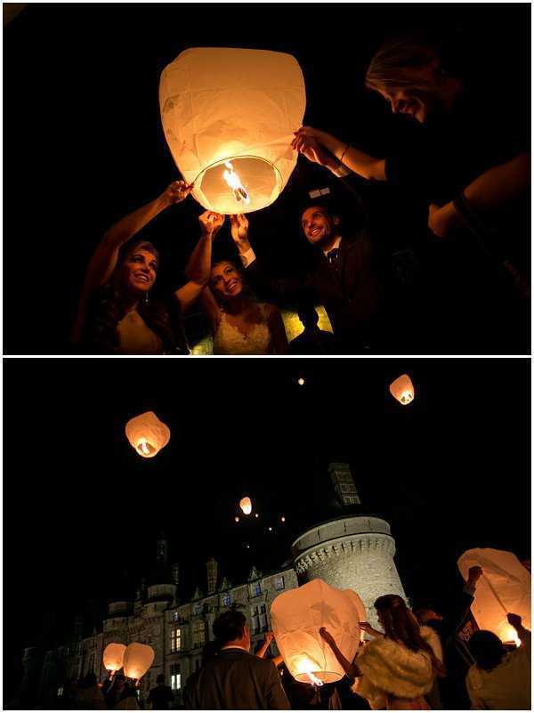 A two-panel nighttime wedding photograph capturing a sky lantern release. The top panel is a close-up portrait showing the bride in a white gown and approximately three other guests — including a man in a dark suit — holding and lighting a white paper sky lantern, their faces warmly illuminated by the orange flame against a completely dark background. The bottom panel is a wide shot showing a group of around ten to fifteen guests standing in front of a large French chateau with a distinctive round tower, each holding glowing white sky lanterns ready for release, with several lanterns already floating into the night sky above the illuminated building. The warm amber glow of the lanterns contrasts sharply with the dark sky and softly lit stone facade of the chateau. Potential venue feature image.