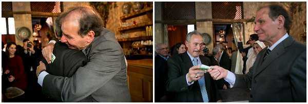 A diptych of two candid reception moments taken indoors in what appears to be a rustic, warmly decorated venue with ornate wooden shelving, traditional lanterns, and ambient lighting visible in the background. On the left, two men in dark suits share an embrace, with a small group of guests visible behind them. On the right, two men in suits — one in charcoal, one in light blue — exchange what appears to be a small green envelope or card, likely a monetary gift, while guests with cameras photograph the moment in the background. Both frames capture close-up, candid guest interaction shots with a warm, low-light indoor atmosphere consistent with a traditional or classic wedding celebration style.