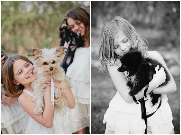 A side-by-side diptych of two portrait shots featuring young flower girls with small dogs at an outdoor wedding. In the left color image, a dark-haired girl in a white ruffled dress laughs while holding a small tan Yorkshire Terrier with a white ribbon; a woman holding a camera is partially visible behind her. The right image is black and white and shows a blonde girl in a white strapless ruffled dress cuddling a small black dog, eyes downcast with a gentle expression. Both portraits are taken in a soft, naturally lit outdoor setting with blurred greenery in the background.