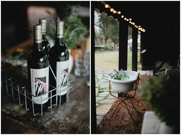 A two-image detail collage showing rustic wedding bar and beverage styling. The left image is a close-up of three dark green wine bottles labeled 'B.R. Cohn' held in a wire bottle carrier on a weathered wooden surface, with greenery visible in the background. The right image shows a white enamel basin filled with ice and bottles, placed on a hay bale atop a stone paver floor, beneath a covered outdoor structure strung with warm globe fairy lights. The overall styling is rustic and casual, with natural and repurposed elements used as decor.