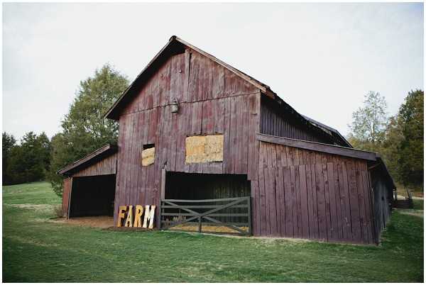 Wide shot of a large rustic red-painted wooden barn with weathered vertical board siding, serving as a wedding venue. Large marquee letters spelling 'FARM' in gold/warm-toned illuminated letters are positioned at the base of the barn's main entrance, which features a wooden gate. The barn has a steep gabled roof and appears to be set on a working farm property. No people are visible in the frame. Potential venue feature image.