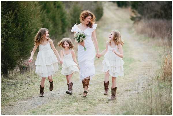 A bride and three flower girls walk together along a rustic dirt path in an outdoor rural setting, all laughing and holding hands. The bride wears a white one-shoulder dress with ruffle detail and brown cowboy boots, carrying a small bouquet of white flowers with greenery. The three flower girls wear tiered ivory dresses with brown cowboy boots, coordinating with the bride's footwear in a rustic country styling theme. The image is a wide portrait shot with a shallow depth of field, capturing a candid, joyful moment.
