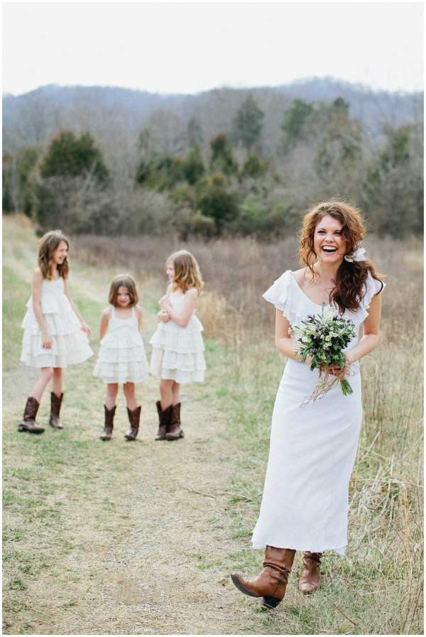 A bride walks along an outdoor dirt path in a rural, open field setting with three young flower girls visible in the background. The bride wears a white midi-length dress with a one-shoulder ruffle detail, paired with brown cowboy boots, and carries a loosely arranged bouquet of greenery and small white wildflowers tied with twine. She is laughing broadly with her hair worn down in loose waves, with a small white flower pinned near her ear. The three flower girls wear white tiered ruffle dresses also paired with brown cowboy boots, consistent with a rustic, country-style wedding theme. The composition is a full-length portrait shot with the flower girls softly out of focus in the background.
