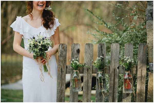 A bride stands outdoors laughing, wearing a white off-shoulder dress with ruffle trim and holding a wildflower bouquet featuring deep purple and white blooms with trailing greenery tied with twine. She is positioned beside a weathered wooden picket fence from which several small glass bottles filled with wildflowers and greenery are hung using twine as rustic decorations. The shot is a medium portrait taken from roughly the waist up, with the bride slightly left of center and the decorated fence occupying the right portion of the frame. The overall styling is rustic-boho with a wildflower and natural twine aesthetic.