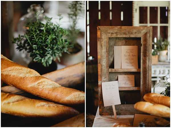 A split close-up detail shot of rustic wedding reception styling. The left panel shows several French baguettes arranged on a surface alongside potted fresh herbs, suggesting a French-inspired food station. The right panel features a weathered wooden shadow-box display holding printed ceremony programs or stationery cards in cream and blush tones, with additional baguettes and greenery visible in the foreground. The overall decor palette is warm and natural, with aged wood, cream paper goods, and fresh herbs contributing to a rustic, farmhouse-style aesthetic.