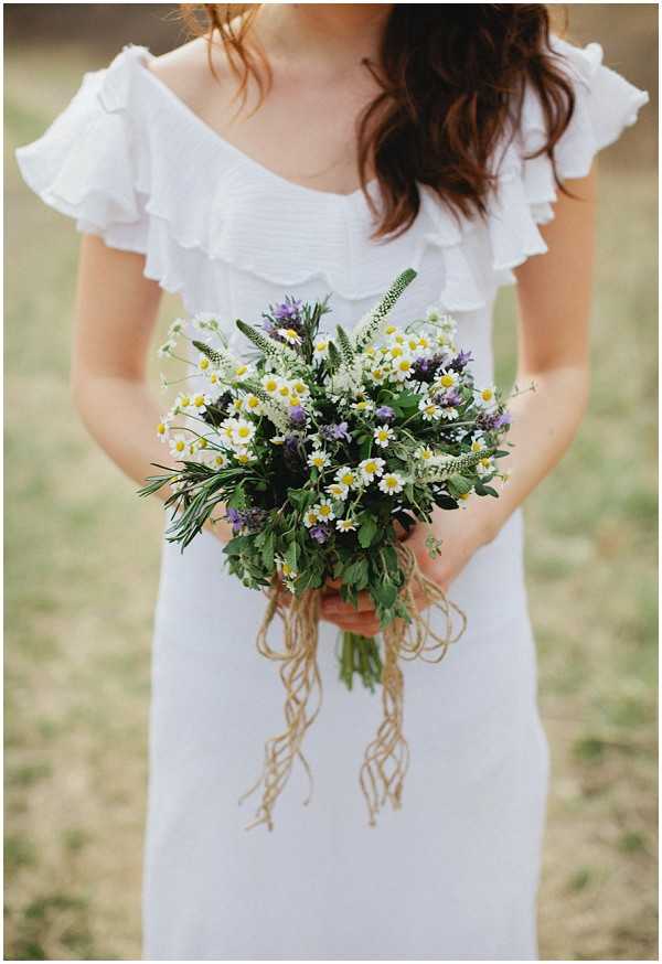 A close-up portrait of a bride from the shoulders down, holding a wildflower bouquet composed of white daisies, small purple flowers, green herbs, and feathery foliage, tied with trailing twine ribbon. The bride wears a white dress with a ruffled, scallop-edged off-the-shoulder neckline and has loose, dark wavy hair. The overall styling is relaxed and bohemian, with the natural, unstructured bouquet reinforcing an informal, garden-gathered aesthetic. The background is softly blurred outdoor greenery.