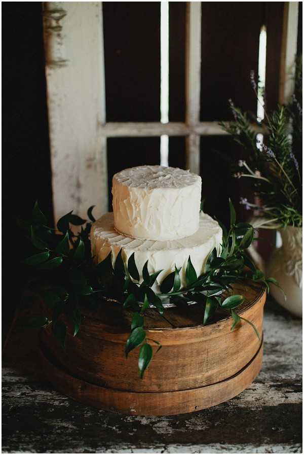 Close-up detail shot of a two-tier wedding cheese wheel cake displayed on stacked rustic wooden circular boards. The two large rounds of cheese are decorated with trailing dark green foliage garlands between and around the tiers. To the right, a cream ceramic vase holds sprigs of lavender and greenery. The display is set in front of a weathered, peeling white-painted window frame in a rustic indoor setting, consistent with a barn or farmhouse aesthetic. The overall styling is rustic and natural, with an earthy, organic decor palette.