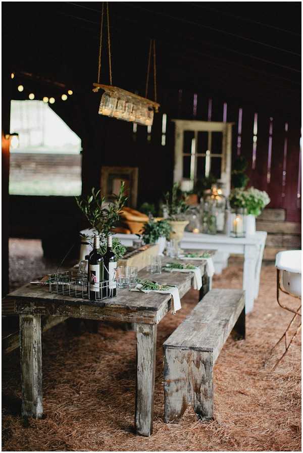 A reception table setup photographed inside a rustic barn venue with a hay-covered floor. The scene features weathered wood farmhouse tables with matching bench seating, styled in an earthy, rustic aesthetic. The tables are decorated with green foliage arrangements, olive branches, glass votives with candles, wine bottles in a wire caddy, and drinking glasses scattered among greenery. In the background, vintage reclaimed window frames are used as decorative props alongside white painted furniture and additional greenery displays. A hanging light fixture made from what appears to be a repurposed wooden or metal frame is suspended from the ceiling by rope, and string lights are visible in the upper background. The overall color palette is muted green, natural wood, and dark barn tones, with no floral color accents beyond greenery. Wide shot with shallow depth of field, taken from a low angle.