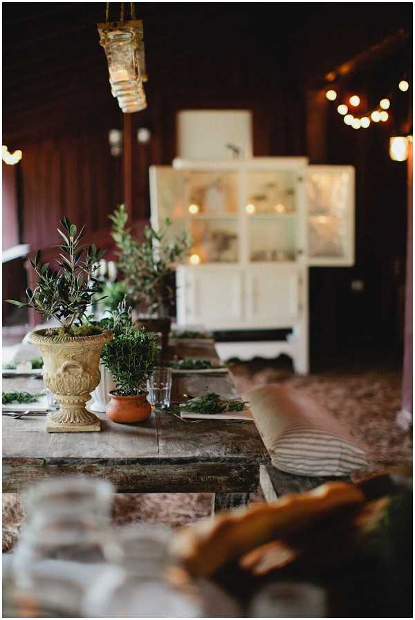 A rustic indoor reception table setup photographed in a medium close-up shot with shallow depth of field. The long wooden farmhouse-style trestle table is decorated with potted herbs and small olive or rosemary plants in terracotta pots and a stone urn planter, along with scattered greenery along the table runner. Striped linen cushions are placed on the bench seating. In the background, a vintage white painted cabinet/armoire is visible, and warm globe string lights are strung across the dark wood-paneled interior space. A hanging glass candle holder is visible in the foreground upper frame. The overall styling is rustic and organic, with an earthy, natural palette of terracotta, green, and natural wood tones.