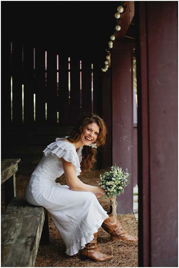 A bridal portrait of a woman seated on a wooden bench inside what appears to be a rustic barn or covered outbuilding, laughing naturally toward the camera. She wears a white midi-length dress with ruffle shoulders and a tiered hem, paired with tan brown leather cowboy-style ankle boots, giving the look a boho-rustic feel. She holds a small, loosely gathered bouquet of white wildflowers, greenery, and small purple accents, tied with twine. A string of round globe lights is visible along the dark wooden beams above her, and hay covers the ground beneath her feet. The portrait is shot from a full-length vertical perspective, framed by the dark barn doorway and a deep red wooden post in the foreground.