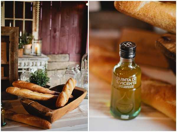 A diptych of close-up detail shots from a wedding reception dinner table. The left image shows a rustic wooden dough bowl filled with small baguettes, with glass vessels, potted rosemary, and a lit pillar candle visible in the background against a distressed red-painted door. The right image is a close-up of a small bottle of Quinta de S. Vicente olive oil placed beside bread rolls, serving as a table favor or condiment. The overall styling is rustic and informal, with natural wood tones and simple, food-focused table details.
