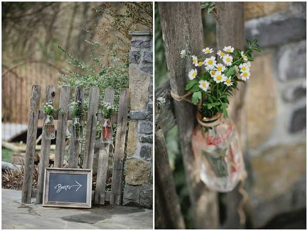 A rustic outdoor wedding detail shot presented as a two-image diptych. The left image shows a weathered wooden picket gate with small glass bottles containing white daisy and greenery arrangements hung from the slats, alongside a chalkboard sign with a handwritten arrow reading 'Barn' directing guests. The right image is a close-up of one of the hanging glass bottles, wrapped in burlap and red-patterned fabric tied with twine, filled with white daisies and green foliage. The overall decor palette is rustic and country-style, using natural textures, twine, and wildflower arrangements consistent with a barn wedding aesthetic.
